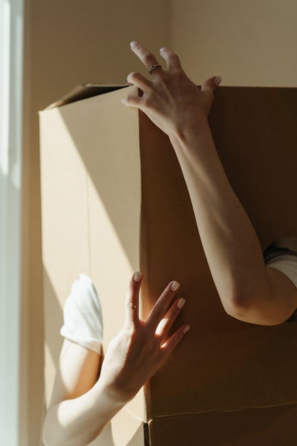 A man with an Afro hairstyle and dark clothing is kneeling on a wooden floor inside a property, surrounded by multiple large and medium-sized cardboard boxes, some labeled with contents such as bedroo