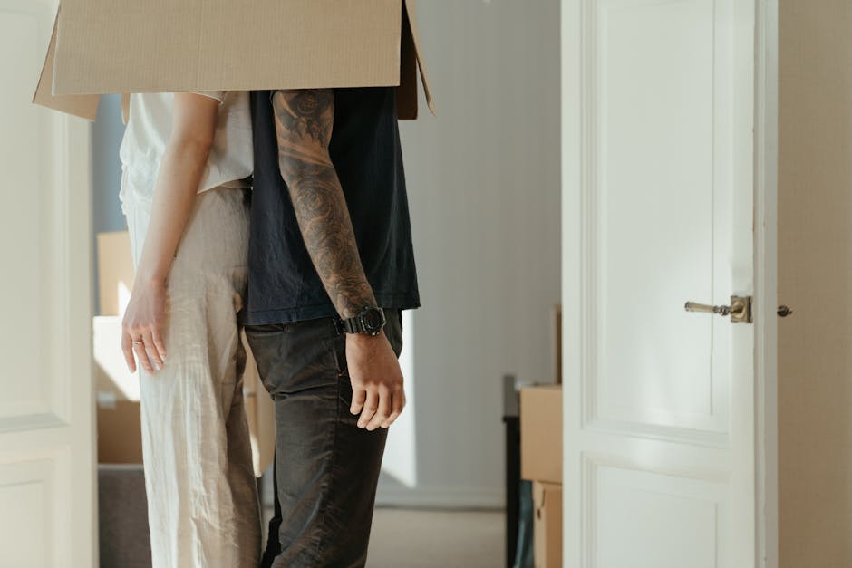 In the image, two movers are seen inside a residential property, preparing for a home relocation. One mover is carrying a large cardboard box, supporting it with both hands, while the other is standing close by, dressed in casual black clothing and wearing a wristwatch. The scene is set in a room with neutral-toned walls and white wooden doorframes, with sunlight filtering through, creating a well-lit environment. In the background, part of a cardboard box and a glimpse of another interior space are visible, indicating an organized packing and moving process. This loading process appears to be taking place near an open doorway leading into a hallway or another room, with various packing materials and possibly furniture or appliances nearby. Such details align with professional removals services provided by Man with Van Bayswater, focusing on efficient furniture transport and packing during house and flat moves in London, including Lancaster Gate and Bayswater areas.