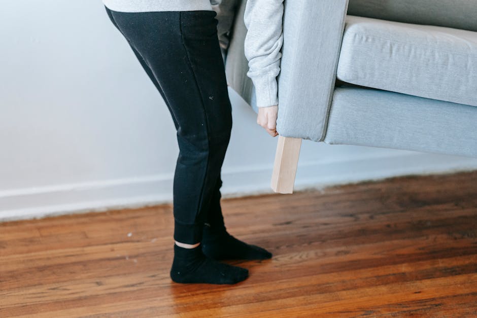 A person wearing dark jeans and black socks is lifting a light grey upholstered sofa with wooden legs, supporting its weight from underneath, inside a home with wooden flooring and a white wall. The sofa appears to be part of a home relocation process, possibly being prepared for transportation or moving through a doorway. The person's hand grips the sofa firmly, demonstrating the careful handling required during packing and moving activities. Visible on the floor are a few small packing materials, indicating that a packing and moving operation is underway. In the background, a white wall and baseboard are visible, suggesting an indoor environment. This scene reflects aspects of furniture transport and loading processes involved in house removals, similar to those handled by companies like Man with Van Bayswater, who offer professional removals services, including lifting and loading furniture during flat moves with lifts, as described in the page about Lancaster Gate removals tips.