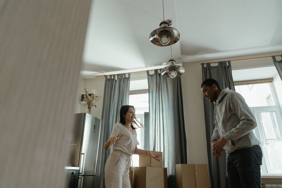 In the image, two movers are seen inside a residential property, preparing for a home relocation. One mover is carrying a large cardboard box, supporting it with both hands, while the other is standing close by, dressed in casual black clothing and wearing a wristwatch. The scene is set in a room with neutral-toned walls and white wooden doorframes, with sunlight filtering through, creating a well-lit environment. In the background, part of a cardboard box and a glimpse of another interior space are visible, indicating an organized packing and moving process. This loading process appears to be taking place near an open doorway leading into a hallway or another room, with various packing materials and possibly furniture or appliances nearby. Such details align with professional removals services provided by Man with Van Bayswater, focusing on efficient furniture transport and packing during house and flat moves in London, including Lancaster Gate and Bayswater areas.