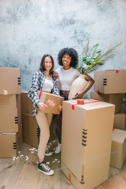 Two women are inside a room with a textured, light-colored wall, surrounded by multiple packed cardboard boxes sealed with red tape, indicating a home relocation or packing process. One woman, wearing
