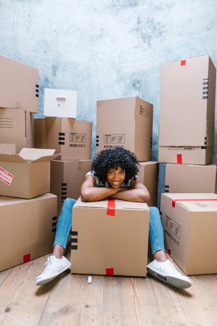Two women are inside a room with a textured, light-colored wall, surrounded by multiple packed cardboard boxes sealed with red tape, indicating a home relocation or packing process. One woman, wearing