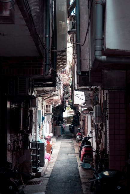 The image depicts a narrow residential alleyway in Bayswater, with tightly packed buildings on either side, featuring visible brick and tiled facades. The alley is cluttered with various belongings such as cardboard boxes, plastic wrapping, and furniture items, some of which are placed on the pavement while others are positioned near doorways and windows. A few small motor scooters are parked along the sides, and there are visible piping and electrical wiring attached to the buildings' exteriors. The scene appears to be in daylight, with natural light illuminating the alley and casting shadows. This setting reflects the typical environment encountered in urban house removals in narrow streets, where careful logistics and space management are essential for successful furniture transport and home relocation. The image is relevant to services provided by Man with Van Bayswater, highlighting challenges in moving activities within constrained street spaces.