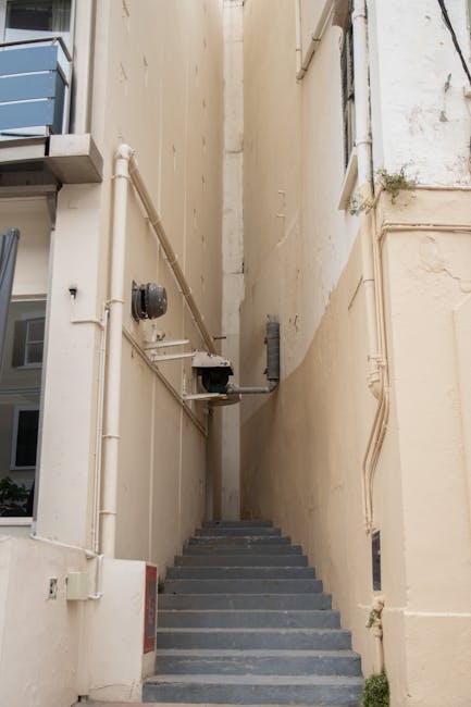 A narrow outdoor alleyway between two tall, cream-colored buildings with visible pipes and electrical conduit running along the walls. Concrete stairs lead upward in the confined space, without any visible handrails. On the left side, part of a blue and gray multi-story building with windows can be seen. The alley appears to be part of a typical urban environment, providing a pathway that may be used during a home relocation or furniture transport process. The lighting is natural, casting soft shadows on the walls and steps. This scene illustrates a typical tight street or alleyway requiring careful navigation during residential moves, with [COMPANY_NAME] often handling such logistics as part of their removal services in Bayswater, especially in areas like Queensway that feature narrow streets and limited access points.